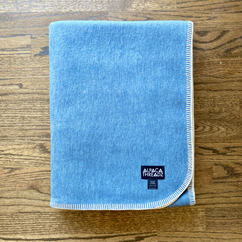 Sky Camp Throw Blanket in soft woven light blue fabric, neatly folded on wooden surface, cozy lightweight design perfect for beach house or cabin décor