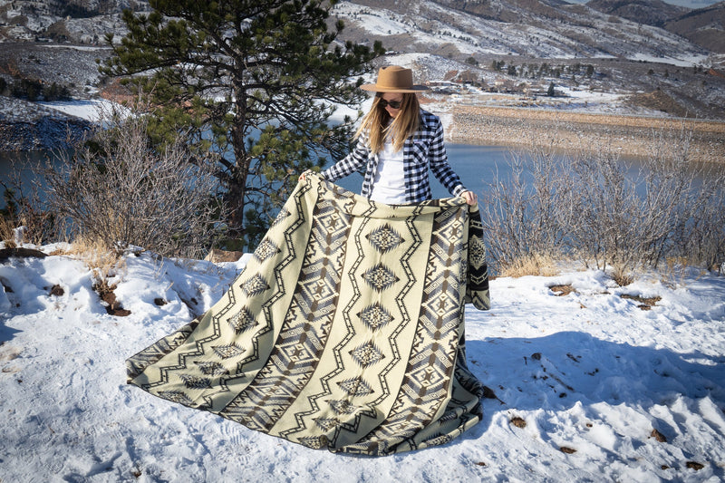 Woman standing on the shore, letting the full length of the Slate alpaca blanket with a geometric pattern drape down into the snow.