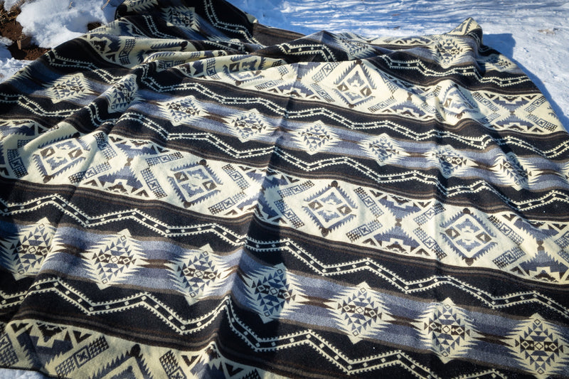 The large Slate alpaca blanket spread fully out on the snow, with sunlight casting shadows that define the geometric weave.