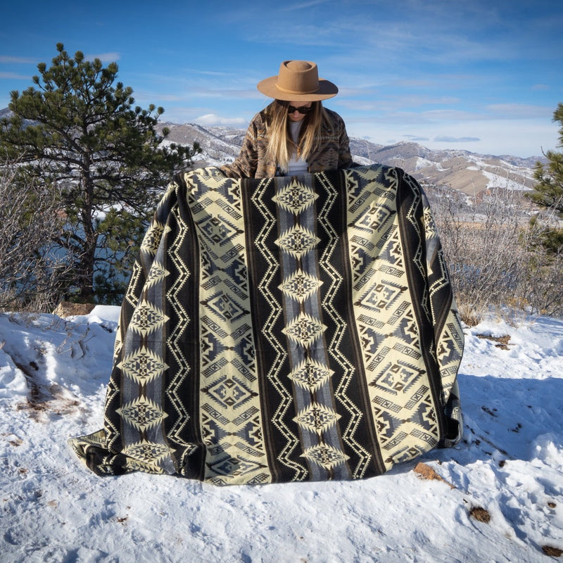 Woman in a hat covered by the Slate alpaca wool blanket with a geometric pattern, standing on a snowy mountain ridge overlooking a valley.
