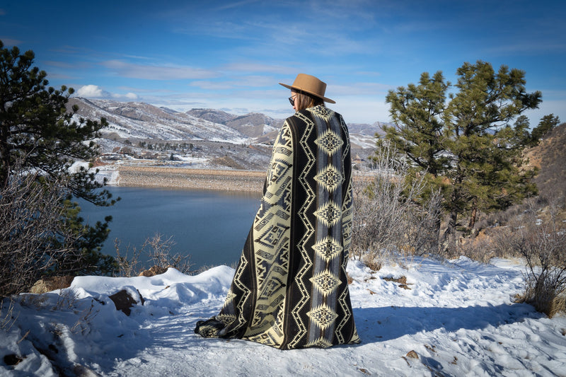 Full body shot of a woman wrapped tightly in the Slate alpaca blanket with a geometric pattern, standing on a snowy hillside by a lake.
