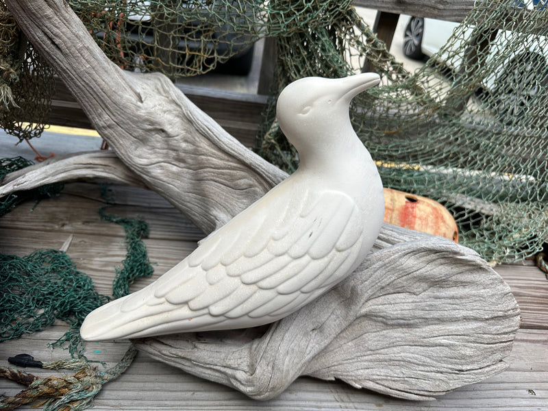 A single white ceramic seagull sculpture, resting on driftwood with a rustic seaside background.