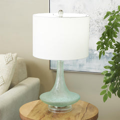 A teal glass table lamp with a white drum shade, placed on a wooden side table in a modern coastal living room.