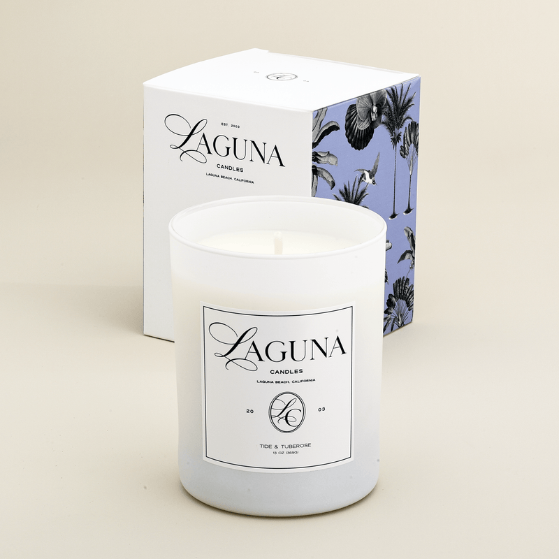 Studio shot of the Laguna Candles Tide & Tuberose 13oz candle next to its white box with lavender and white tropical palm illustration.