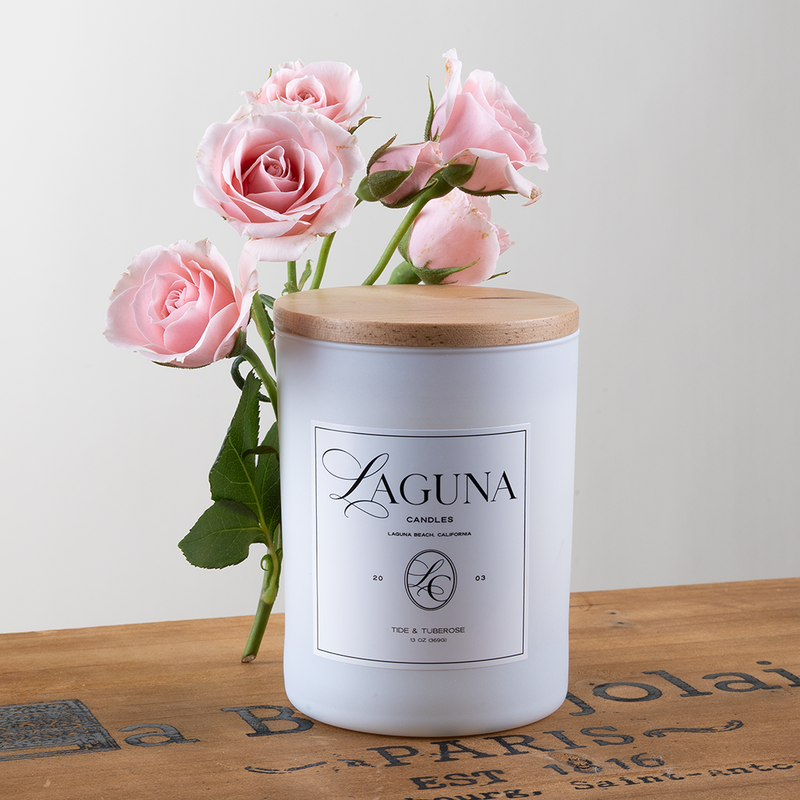 Styled shot of the Tide & Tuberose 13oz candle with a natural wood lid, accented by fresh pink roses and a rustic wooden background, emphasizing the rich floral scent.