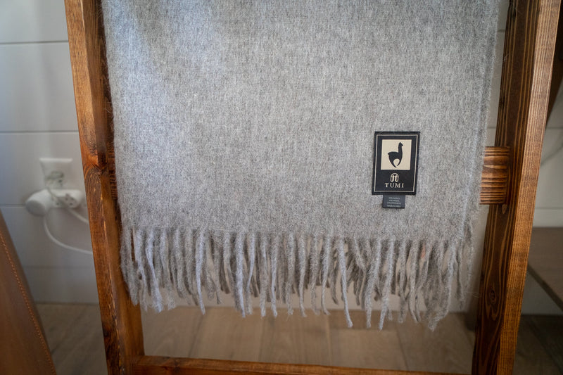 Gray Tumi Alpaca Wool Throw Blanket displayed on a rustic wooden ladder, showcasing soft textured fabric, long fringe edges, and the signature brand label.