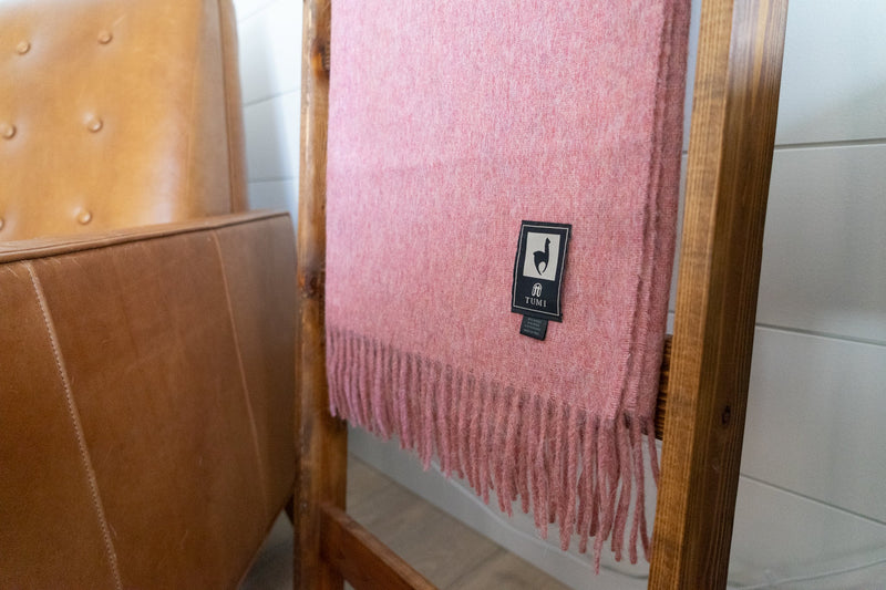 Pink Tumi Alpaca Wool Throw Blanket draped on a wooden ladder beside a tan leather chair, featuring soft fringe edges and the woven brand label.