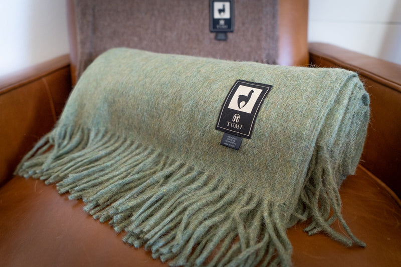 Soft green Tumi Alpaca Wool Throw Blanket neatly rolled on a tan leather chair, showing woven label and long fringe edges with a brown blanket in the background.