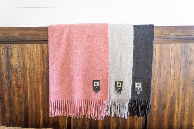 Three Tumi Alpaca Wool Throw Blankets in soft pink, light gray, and black hanging side by side against a rustic wooden background, each featuring fringe edges and the woven Tumi brand label.