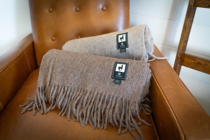 Two Tumi Alpaca Wool Throw Blankets in warm brown and soft cream tones resting on a tan leather chair, showcasing their fringe detailing and woven brand labels.