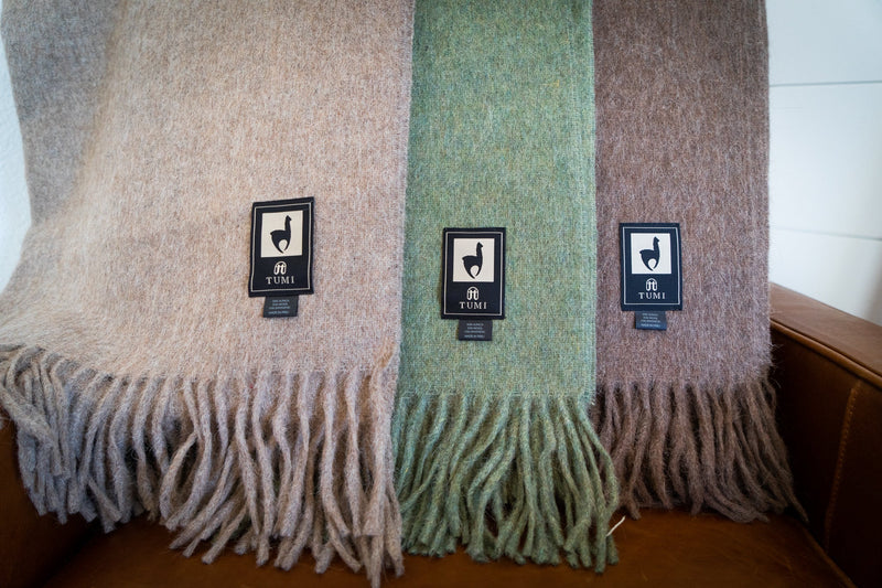 Three Tumi Alpaca Wool Throw Blankets in soft beige, sage green, and cocoa brown draped on a tan leather chair, featuring fringe edges and signature woven brand labels.