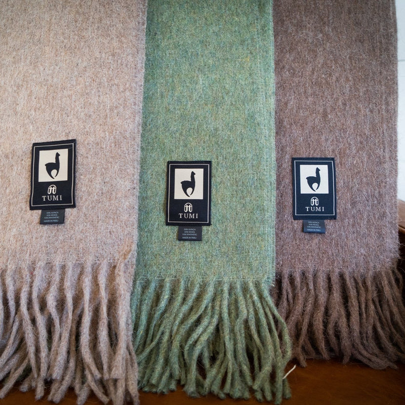 Three Tumi Alpaca Wool Throw Blankets displayed side by side in soft neutral tones of beige, green, and brown with fringe edges and woven labels, set on a wooden surface.