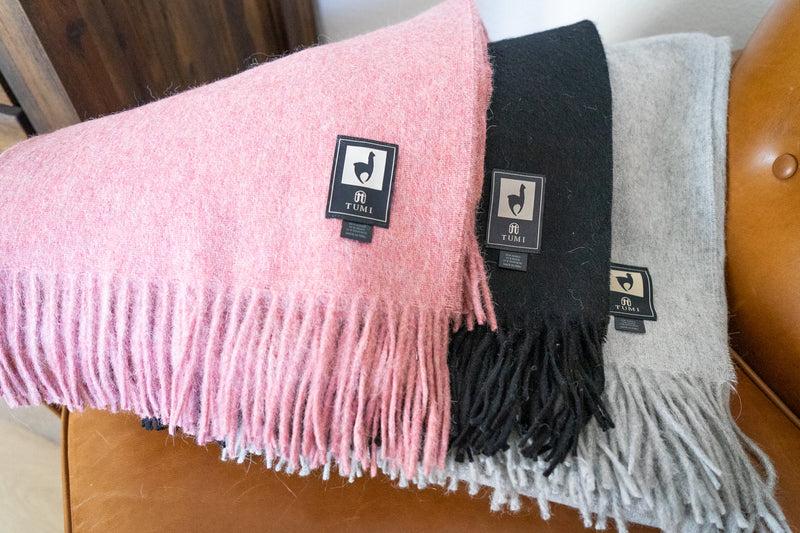 Three Tumi Alpaca Wool Throw Blankets in soft pink, black, and gray displayed on a tan leather chair, showing woven brand labels and fringed edges.