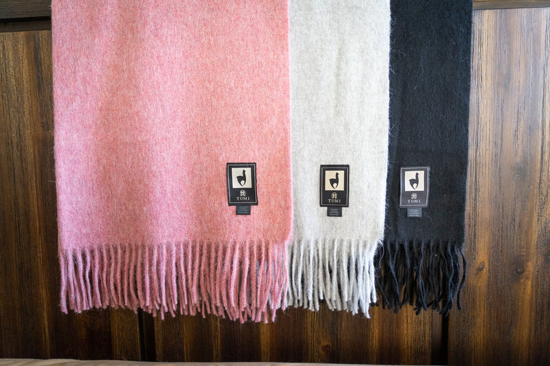Three Tumi Alpaca Wool Throw Blankets in blush pink, light gray, and black hanging neatly, each with soft fringe edges and signature woven brand tags.