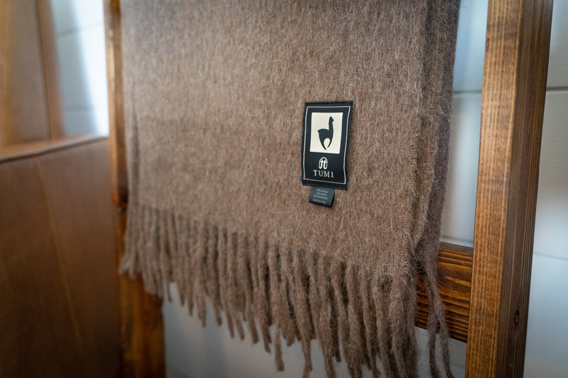 Close-up of the brown Tumi Alpaca Wool Throw Blanket showing the woven texture, fringe detail, and signature Tumi brand label on a wooden ladder.