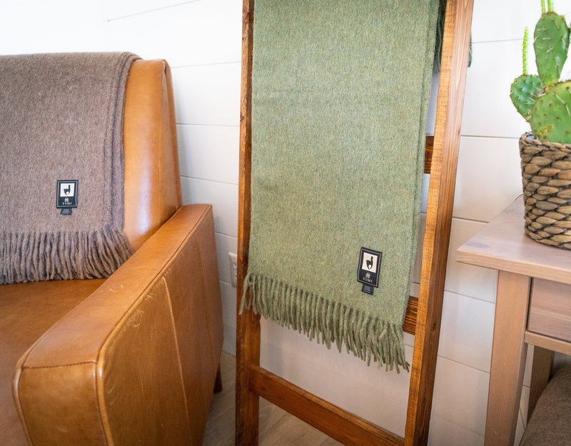 Two Tumi Alpaca Wool Throw Blankets displayed in a cozy corner, one in sage green hanging from a wooden ladder and the other in warm brown draped over a leather chair beside a small table with a cactus plant.