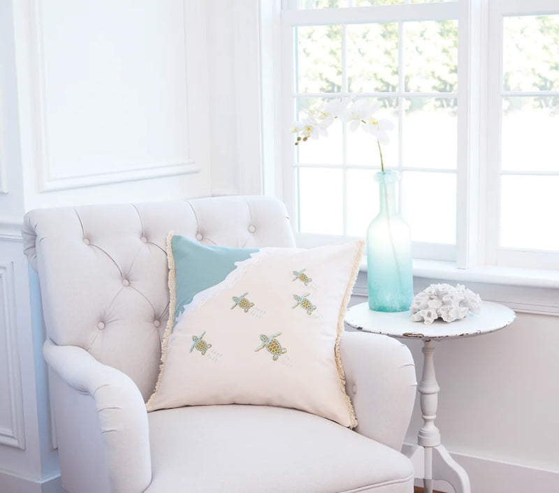 Turtle Migration Indoor Cotton Pillow featuring embroidered baby turtles, fringed edges, and a cream and sea foam design, displayed on an accent chair.