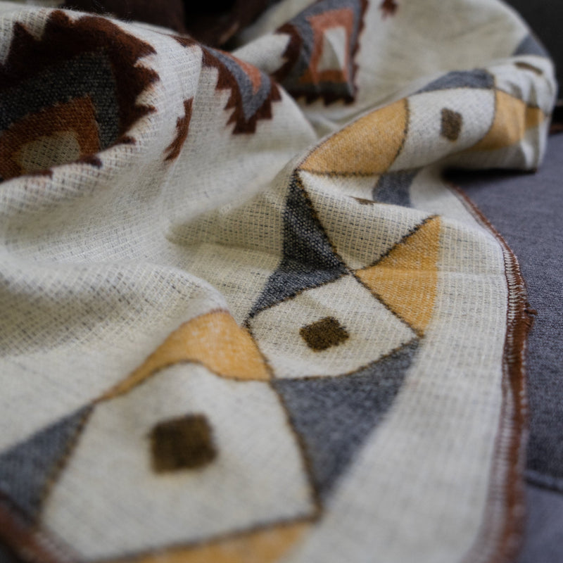 Close-up of a cream-colored Andean Alpaca Wool Throw Blanket, folded, showing a geometric pattern in golden yellow, soft gray, and rust-brown accents on a neutral background.