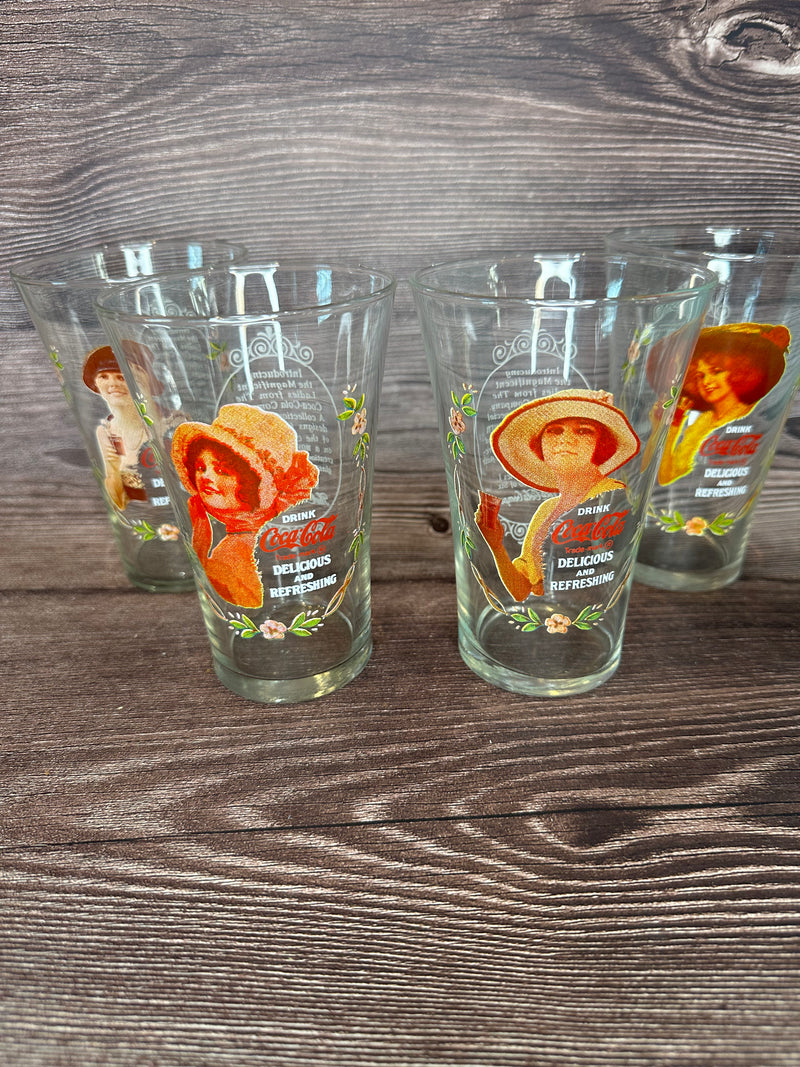 Vintage Coca Cola Magnificent Ladies Collector Series 1 Glasses featuring vibrant images of women in classic Coca-Cola advertising, displayed in a set of four on a wooden surface.
