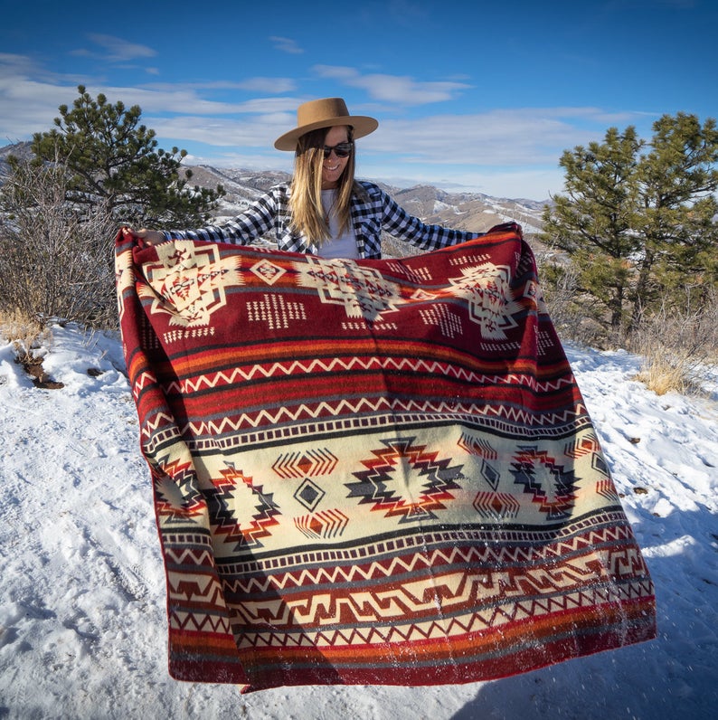 Woman holding the Wildfire blanket draped over one shoulder, highlighting the bold red and geometric motifs against the mountain backdrop.