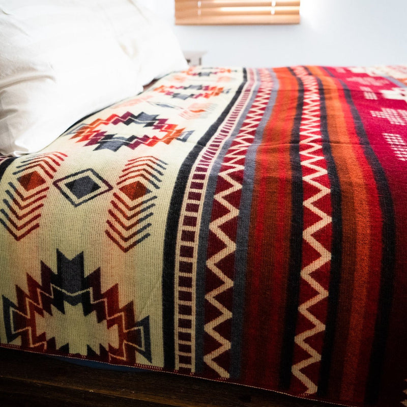 Wildfire Alpaca Blanket draped over a dark wooden headboard, showing the red, cream, and geometric design in a bedroom setting.