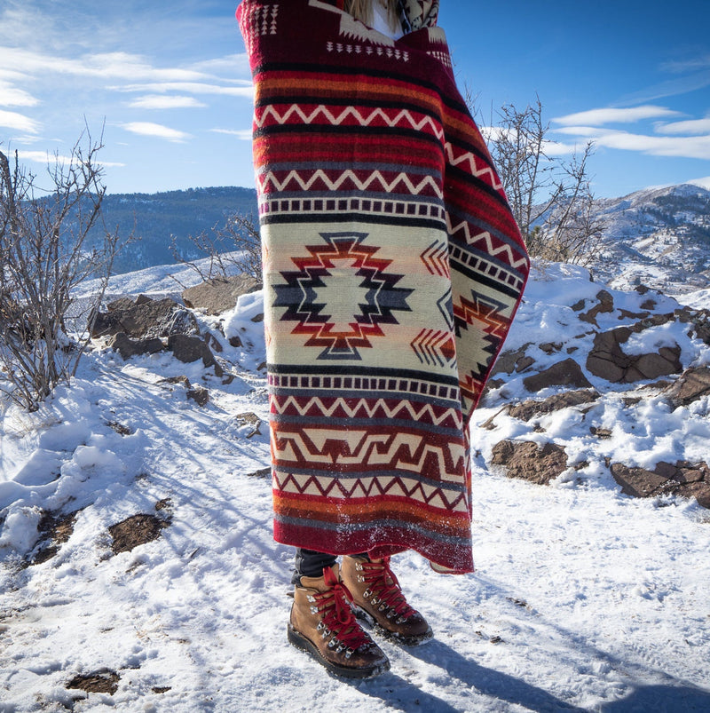 Woman's lower body and boots wrapped in the Wildfire Alpaca Blanket, showcasing the red, burgundy, and cream geometric pattern on a snowy trail.