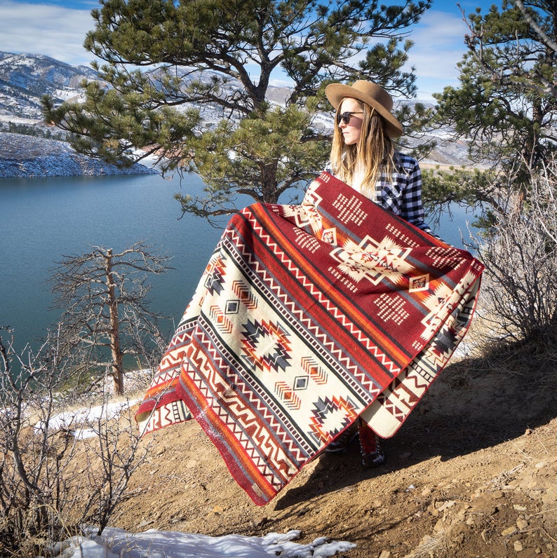 Woman wrapped in the Wildfire Alpaca Blanket, viewed from a side profile in a scenic, golden field with a lake in the background.