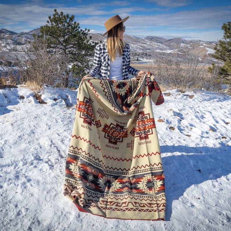 Woman fully wrapped in the Wildfire Queen Alpaca Blanket, standing on a snow-covered hillside with a scenic view behind her.