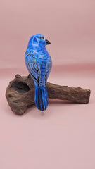 Hand-carved and painted blue bird sculpture perched on a natural driftwood base against a pink background.