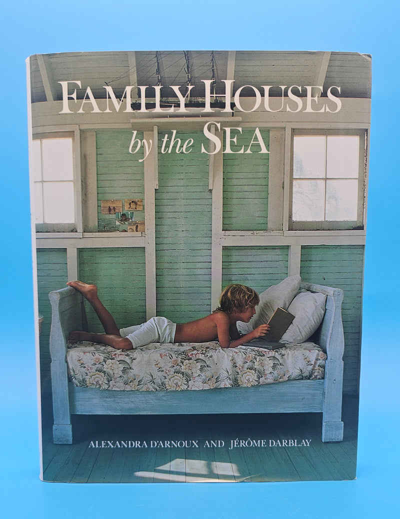 Hardcover of Family Houses by the Sea showing a child reading on a floral daybed inside a green-painted seaside cottage with white trim and sunlit windows.
