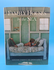 Hardcover of Family Houses by the Sea showing a child reading on a floral daybed inside a green-painted seaside cottage with white trim and sunlit windows.