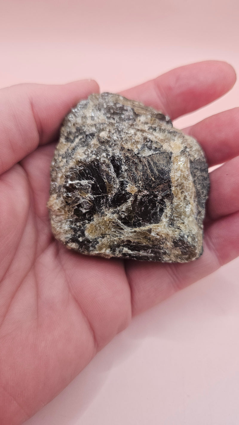 Close-up of a 106g Madagascar Labradorite Rough Specimen with natural gray matrix and subtle flashes of iridescent blue and green, displayed on a light pink background.