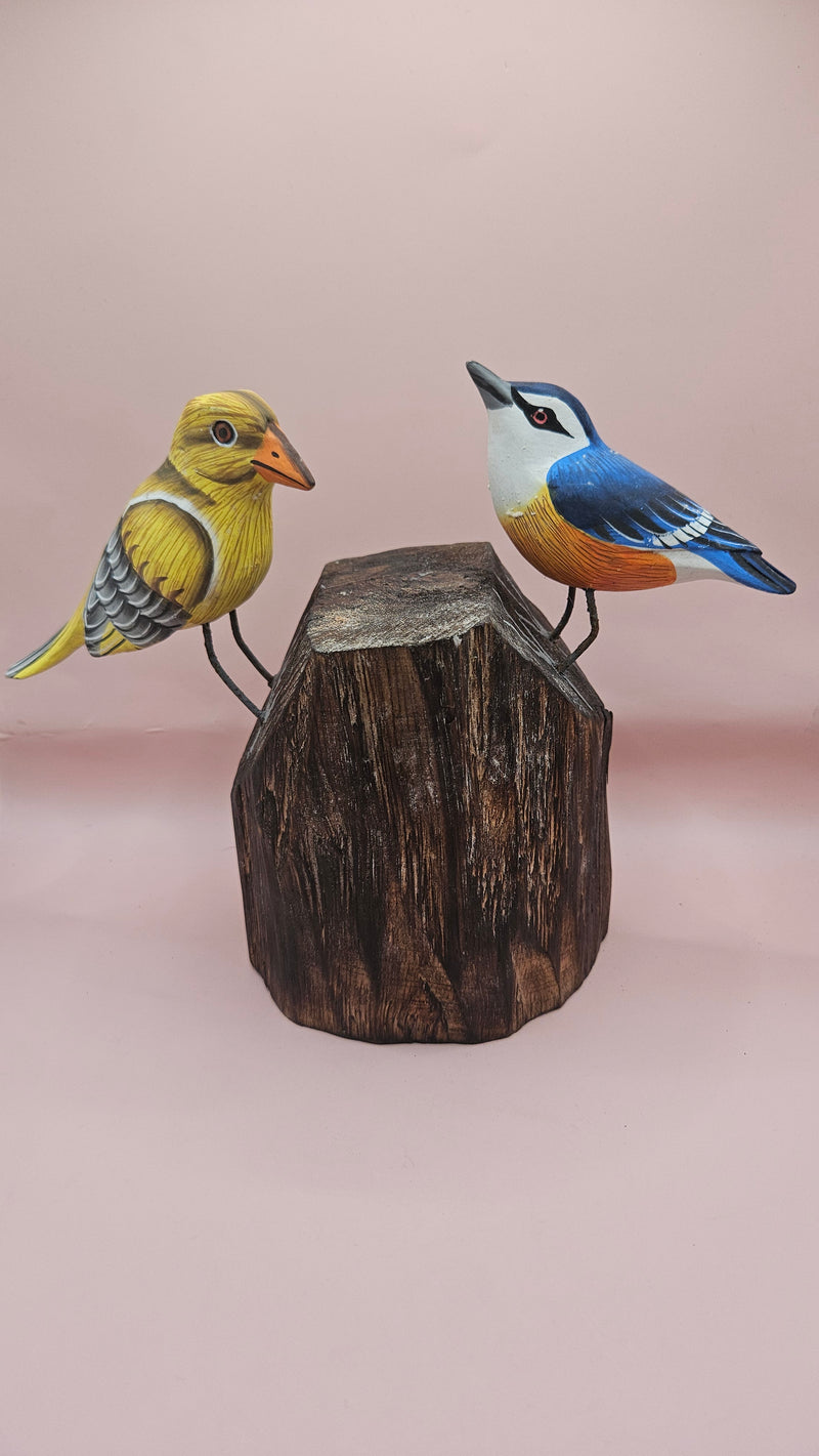 Hand-painted wooden songbird figurines in red/yellow and blue/yellow perched on rustic wood stumps, displayed on a green tabletop.