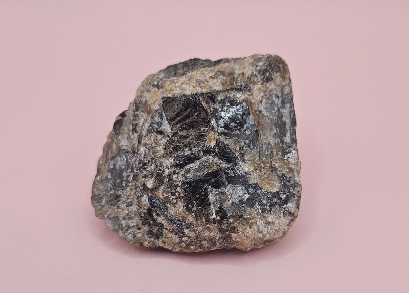 Close-up of a 106g Madagascar Labradorite Rough Specimen with natural gray matrix and subtle flashes of iridescent blue and green, displayed on a light pink background.