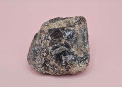 Close-up of a 106g Madagascar Labradorite Rough Specimen with natural gray matrix and subtle flashes of iridescent blue and green, displayed on a light pink background.