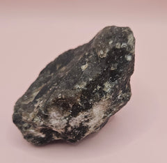 Close-up of a 270g Madagascar Labradorite Rough Specimen with natural gray matrix and subtle flashes of iridescent blue and green, displayed on a light pink background.