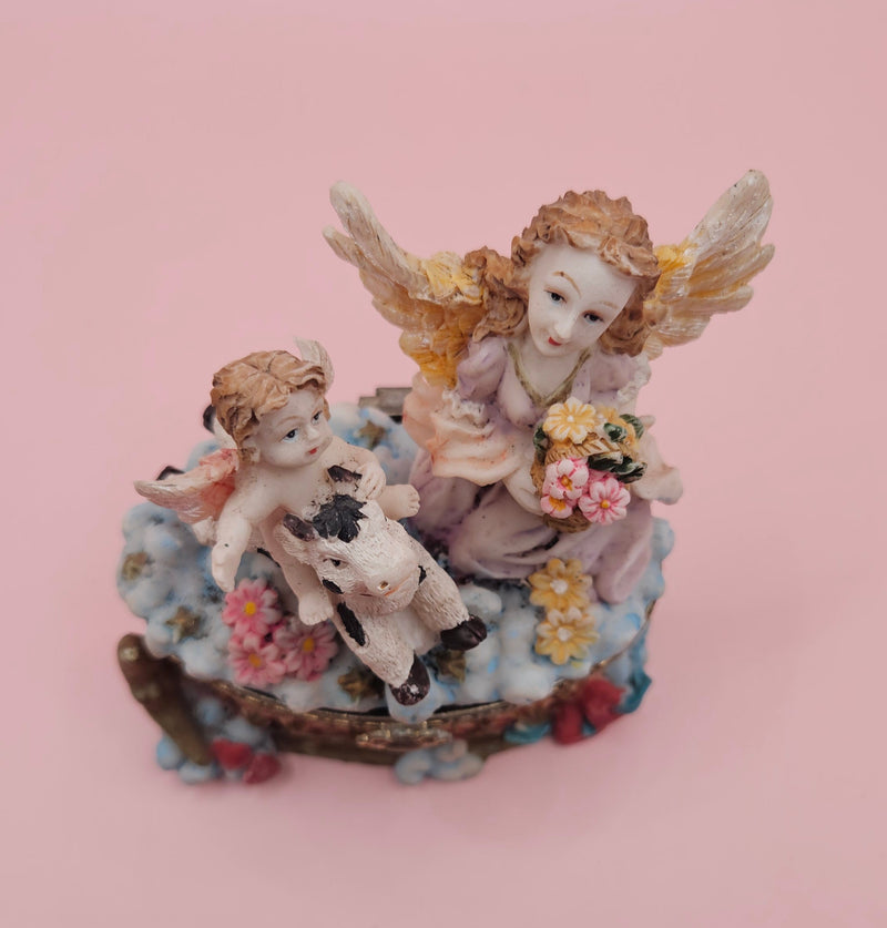 Whimsical vintage trinket box featuring a winged angel holding a basket of flowers, a cherub riding a spotted cow, and decorative star details on a round pastel base.