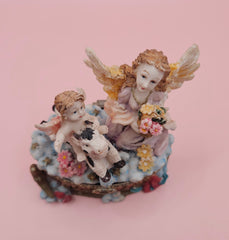 Whimsical vintage trinket box featuring a winged angel holding a basket of flowers, a cherub riding a spotted cow, and decorative star details on a round pastel base.