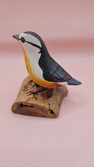 Photo of hand-carved wooden songbird figurine with grey, white, and yellow painted details, perched on a natural driftwood base.