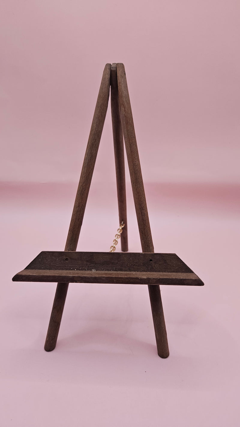 Rustic mini wood easel with dark shelf, set against a pink background. Vintage-style handmade display stand.