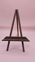 Rustic mini wood easel with dark shelf, set against a pink background. Vintage-style handmade display stand.