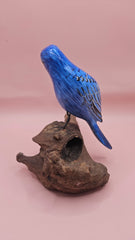 Hand-carved and painted blue bird sculpture perched on a natural driftwood base against a pink background.