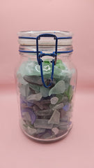 Clamp-lid glass jar with blue hinge filled with multicolored sea glass in green, blue, and frosted white, coastal home decor on pink background