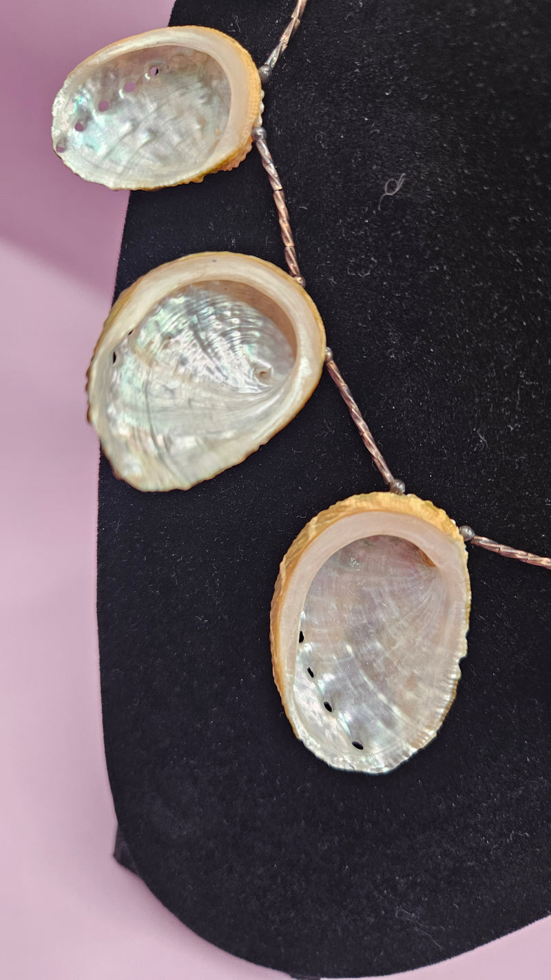 12.25" vintage abalone shell necklace with iridescent polished shells on a delicate chain, displayed on a black stand with a soft pink background.