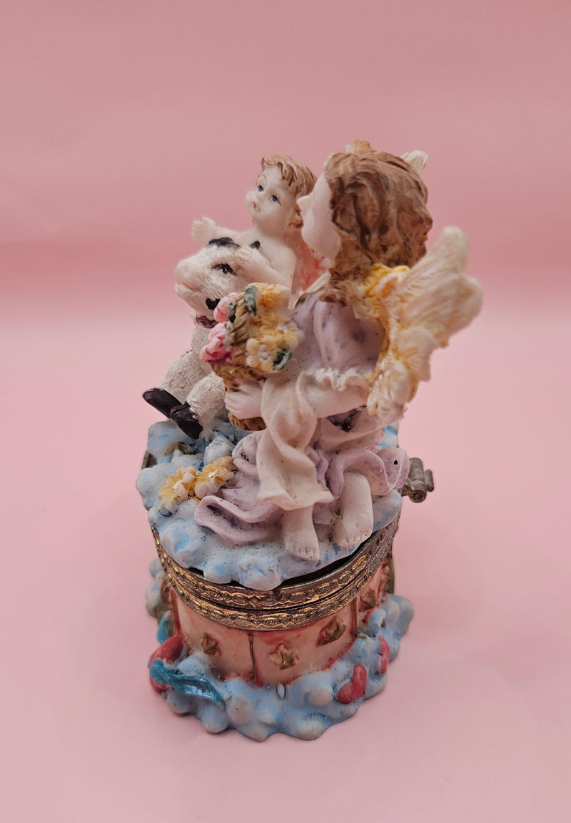 Whimsical vintage trinket box featuring a winged angel holding a basket of flowers, a cherub riding a spotted cow, and decorative star details on a round pastel base.