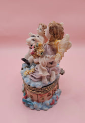 Whimsical vintage trinket box featuring a winged angel holding a basket of flowers, a cherub riding a spotted cow, and decorative star details on a round pastel base.