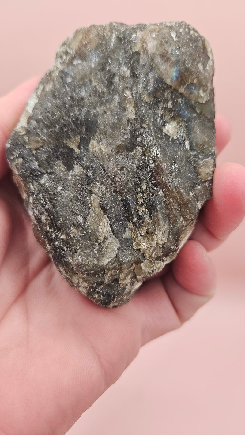 Close-up of a 270g Madagascar Labradorite Rough Specimen with natural gray matrix and subtle flashes of iridescent blue and green, displayed on a light pink background.