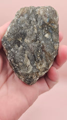 Close-up of a 270g Madagascar Labradorite Rough Specimen with natural gray matrix and subtle flashes of iridescent blue and green, displayed on a light pink background.