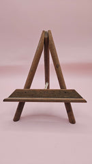 Rustic mini wood easel with dark shelf, set against a pink background. Vintage-style handmade display stand.