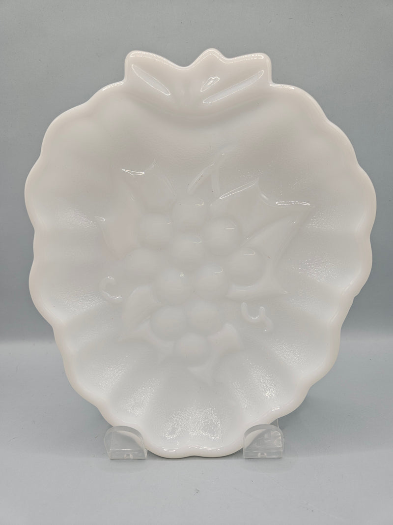 A close-up view of a vintage milk glass plate with a detailed embossed grapevine design. The pristine white plate measures 7" x 5.5" and adds a classic touch to any home decor setting